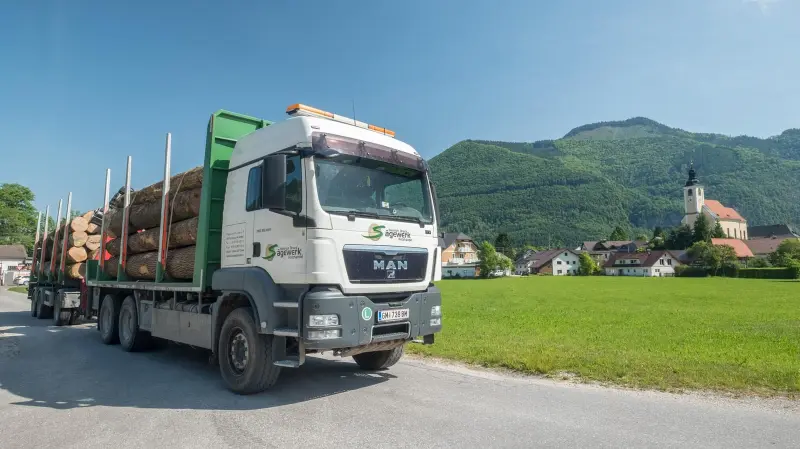 Holztransporter des Sägewerks mit Rundholz vor dem Almtal mit Kirche und Bergen