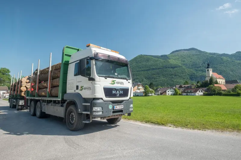 Holztransporter des Sägewerks mit Rundholz vor dem Almtal mit Kirche und Bergen